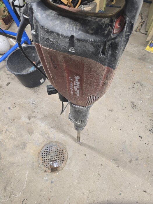 Hilti-bilningsmaskin ovanför en golvbrunn på ett betonggolv i ett garage, med verktyg och hink i bakgrunden.