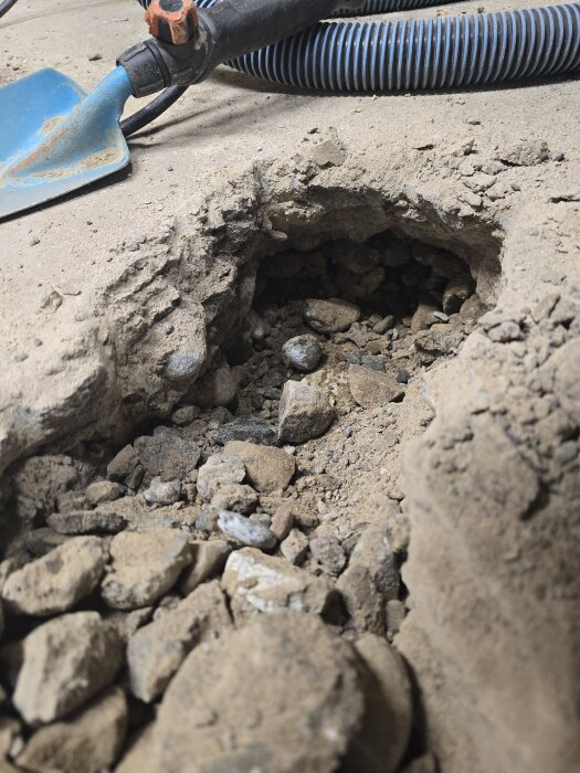 Hål i betonggolv med grus och sand i botten, en liten spade och ett blått plaströr syns ovanpå golvet.