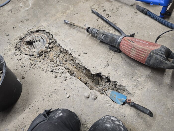 Betonggolv under renovering med golvbrunn synlig, en borrliknande maskin och en liten trädgårdsspade ligger bredvid det uppgrävda avloppsröret.