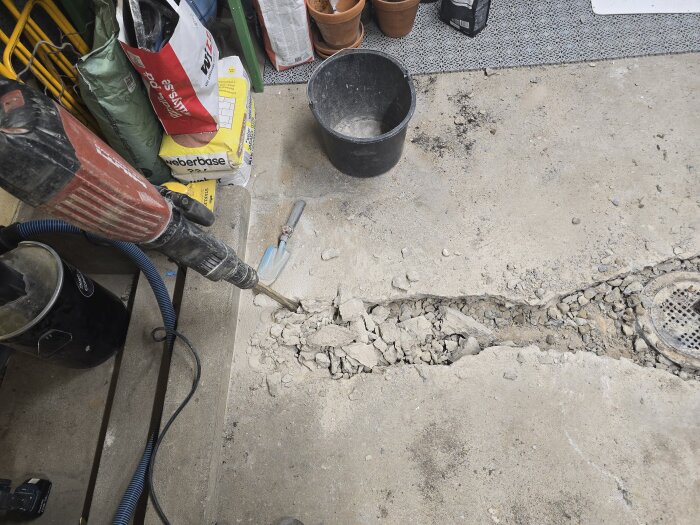 Betonggolv i ett garage bryts upp med en borrhammare. En trädgårdsspade, hink och byggmaterial syns. Avloppsröret är delvis exponerat.