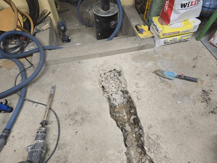 Borrhammare, trädgårdsspade och borrhål i ett betonggolv, omgivet av verktyg och byggmaterial.