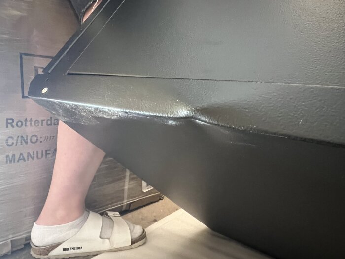 Deformerat nätverksrack i färgen svart med en buckla på sidan, med en person i vita sandaler bredvid.
