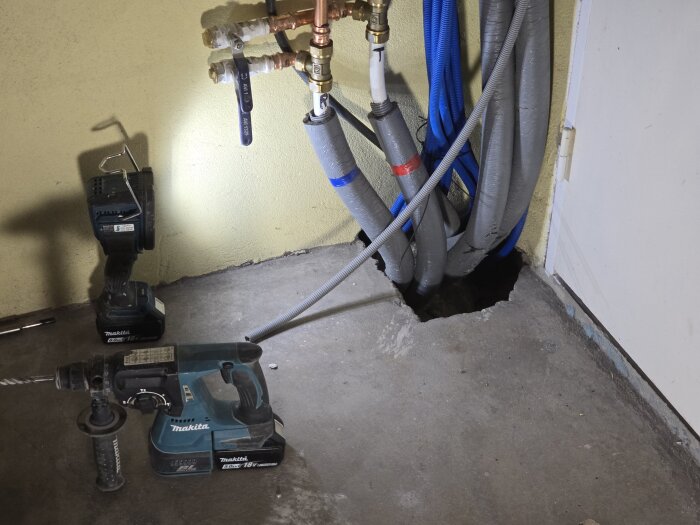 En borrmaskin och rör kopplade till väggen vid ett hål i golvet, med inplastade ventilrör och en sladd i fokus.