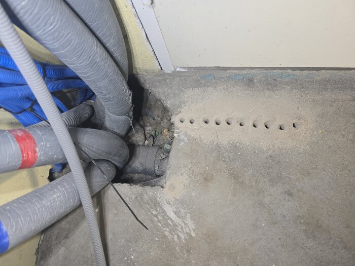 Hål tagna i betong för avloppsrörsdragning, omgivna av grå och blå kablar vid vad som verkar vara en väggnära plats.