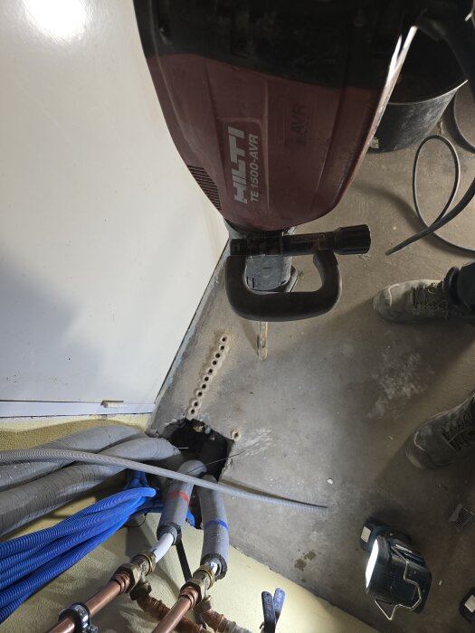 Hilti-maskin över hål i betonggolv med rör och kablar synliga, använd för att bila upp gammalt avloppssystem.
