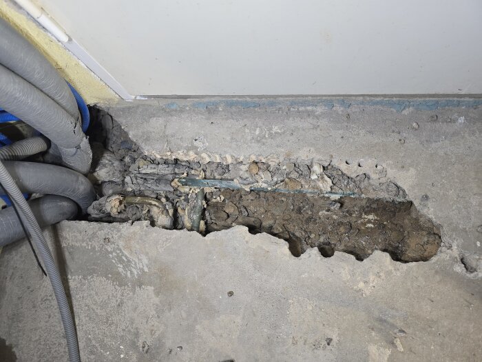 Betonggolv uppbrutet med synliga avloppsrör och kablage, delvis täckt av jord och smuts, under renovering i garage.