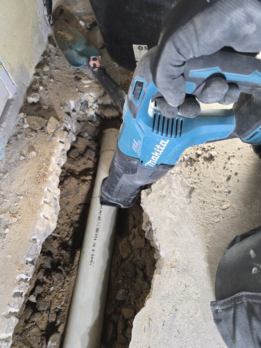 En person använder en blå borr från Makita för att frilägga ett vitt avloppsrör i en uppbruten cementyta.