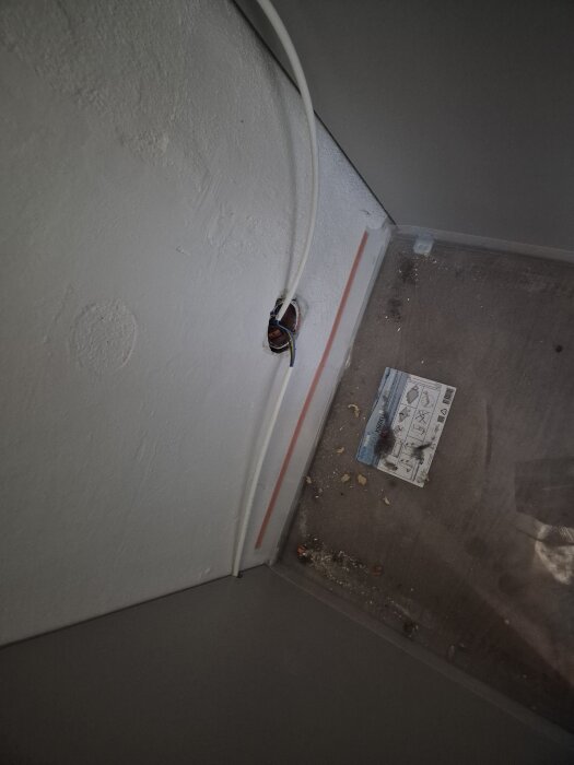 Kabel bakom kylskåp som går genom en vägg, med en täckskiva och golvbrädor. Spår av sågspån och en oskyddad elkabel synlig.