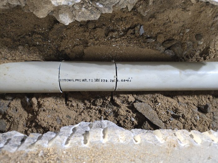 Ett PVC-avloppsrör i en uppborrad ränna i golvet, omgivet av sand och sten, med synlig tryckt text på röret.