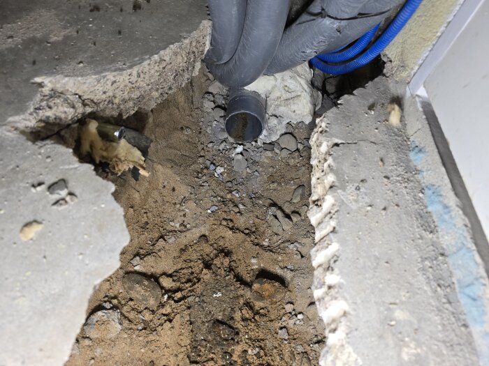 Öppet betonggolv med synliga rör och grus, där avloppssystemet håller på att tas bort och ersättas, visar byggprocessens framsteg.
