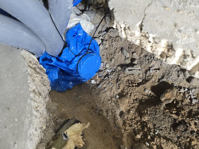 Bilden visar en del av en uppbruten golvsektion med synliga rör, varav ett är täckt med en blå plastpåse och buntband, omgivet av jord och betongrester.