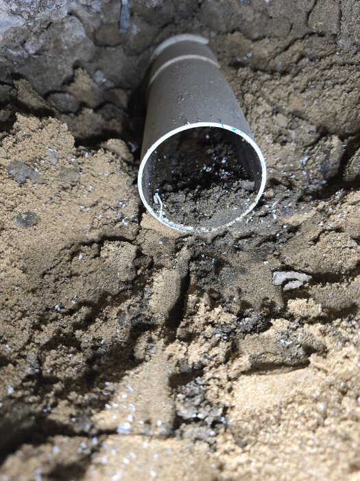 Ett rör av PVC ligger delvis utgrävt i jord, med smuts och skräp som fyller röröppningen.
