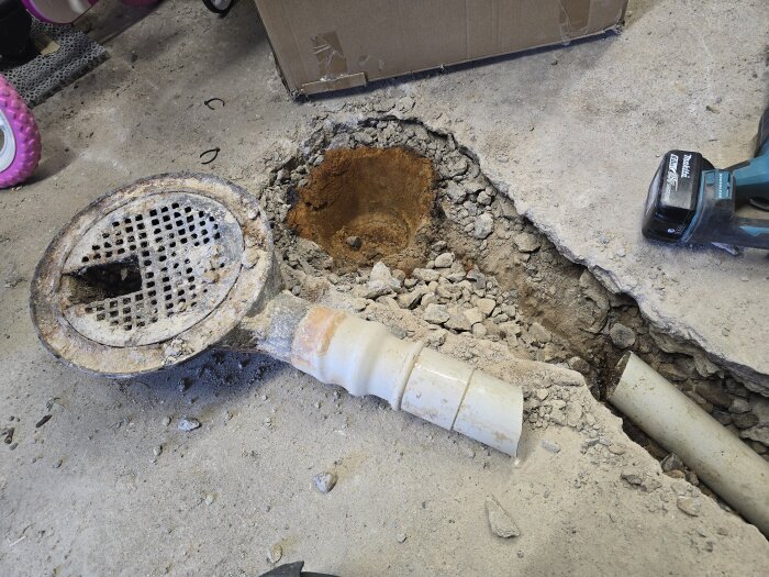 Rostig gjutjärnsbrunn och avloppsrör på sprucken betonggolv uppbilad under renovering, med en borrmaskin intill.