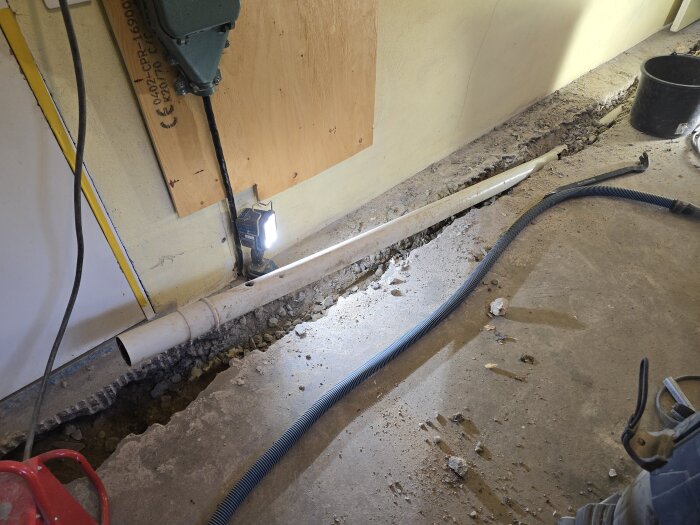 Uppbruten betonggolv med synligt avloppsrör, borr, ficklampa och verktyg. Byggarbete pågår i ett garage.