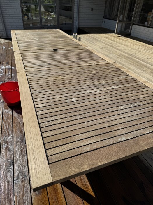 Teakbordsskiva, nyligen rengjord och i solljus, med intilliggande ljusbrun altan, röd hink vid sidan.