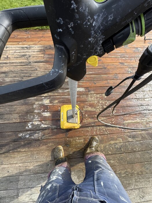 En person rengör en smutsig altan med en Kärcher PCL 6 altantvätt. Maskinen ses ovanifrån, och personen har fläckiga kläder och skor.