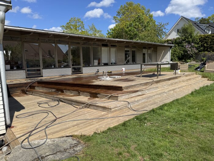 Altan i trä under renovering, delvis rengjord med rengöringsmaskin. Bord och utrustning synliga, solig dag med grönska och hus i bakgrunden.
