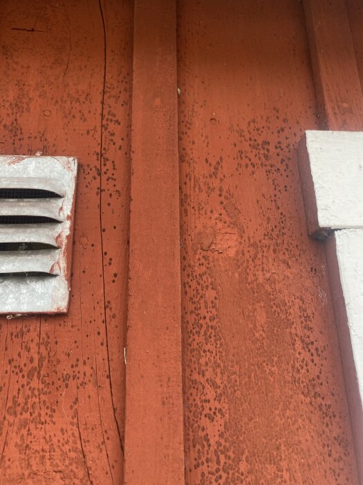 Röd träfasad med mörka prickar, misstänkt mögel, intill vitmålad kant och ventil.