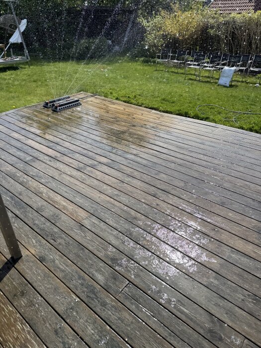 Solbelyst altan delvis blöt efter rengöring, en vattenspridare syns i bakgrunden, och trädgårdsmöbler står i gräset.