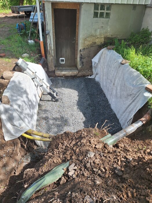 Grävt hål framför källardörr med grusbotten, vit geotextil på sidorna och dräneringsrör, preparerat för stödmurar och trappa.