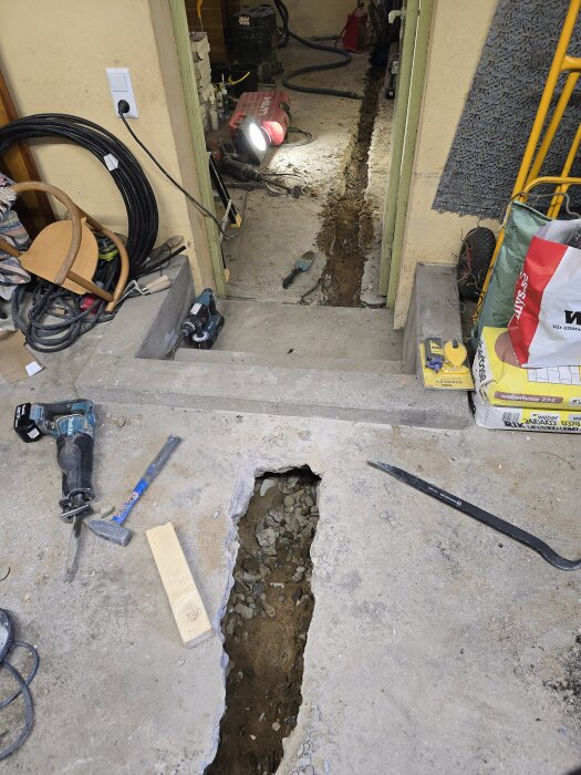 Rivna golvbrunnar med verktyg och en djup grävad skåra i betonggolv, förberedelse för nya avloppsrör i husrenovering.