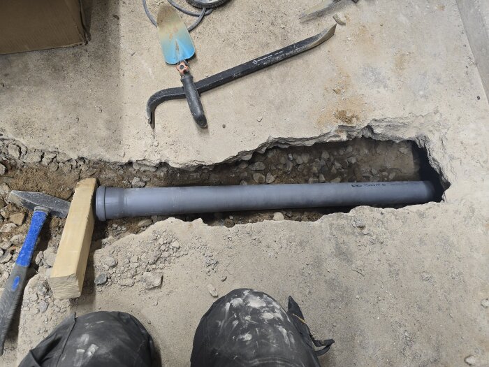 Plaströr installerat i öppet golvhål med verktyg och trästöd runt om. Grus och betongrester syns runtom röröppningen.