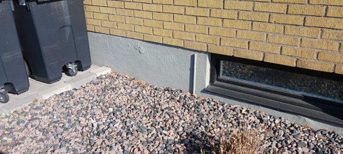 Utomhusvägg av tegel med behov av målning, grus vid foten och en liten källarglugg, samt två svarta soptunnor på hjul till vänster.