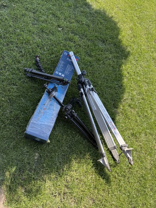 Stativ och förpackning ligger på gräs. Två stativ av metall och plast syns, ena märkt Bosch.