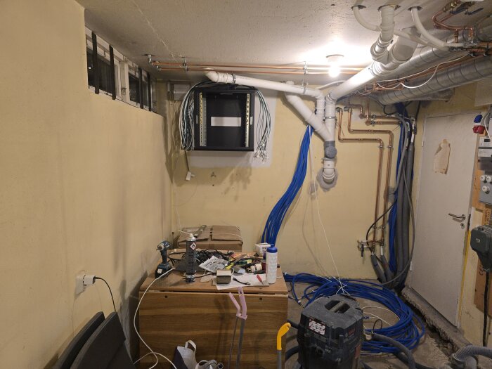 Källare under renovering med öppet nätverksrack, oordnade kablar och rörledningar längs väggen. Verktyg och utrustning på ett bord.
