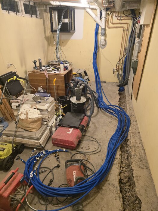 Rum med byggställning, blå kablar spridda över golvet, verktyg och utrustning. Avloppsrör och elinstallation synliga längs väggarna.