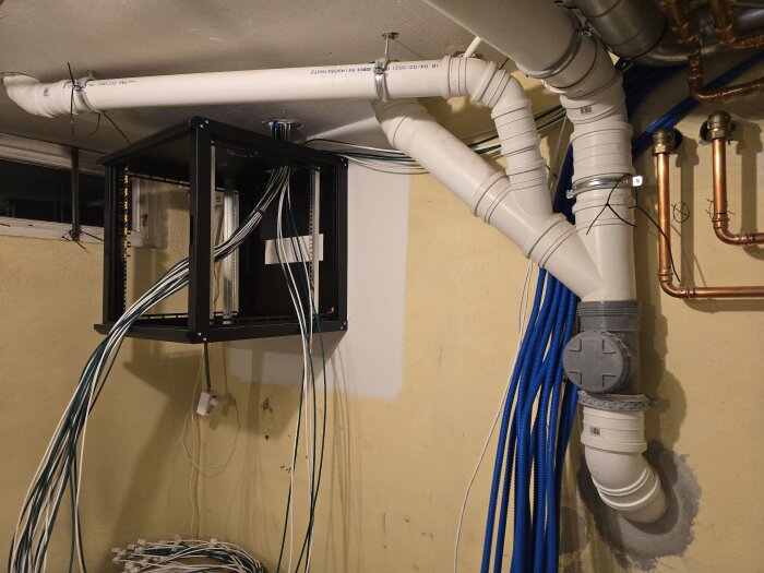 Väggmonterat nätverksrack med kablar och VVS-rör; nätverksinstallation under arbete.