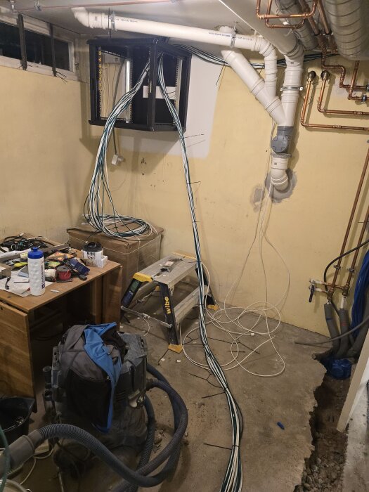 Nätverksrummel med kablar hängande från racket på väggen, arbetsbord med verktyg, stege och dammsugare i rörigt rum under renovering.
