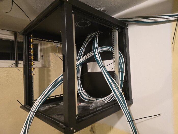 Öppnat nätverksrack på vägg, fullt med osorterade kablar i blå och vita slangar, redo för organisering och installation.