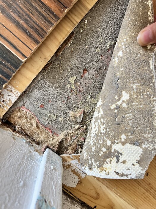 Nedsliten plastmatta med synlig juteväv under lager av parkett och gammalt golv. Potentiell asbest i golvmaterialet diskuteras.