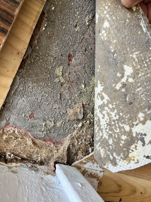 Gammal golvmatta med sliten yta, delvis upplyft för att visa ett lager av damm och smuts, och exponerar gammal jutevävslag med hårstrån.