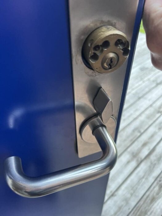 Grått dörrhandtag och lås på en blå dörr, saknar synliga skruvar för att ta av handtaget.