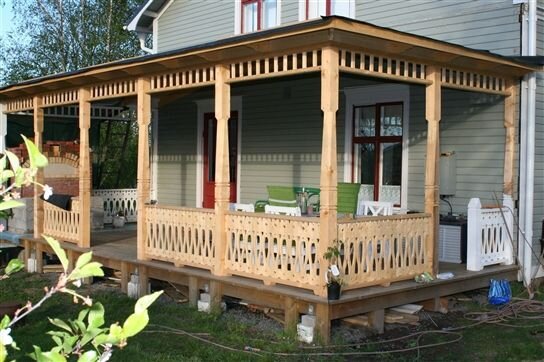 Altan med tak i trä ansluten till hus, dekorerad med räcken och pelare. Möbelgrupp i grönt och vitt syns på altanen.