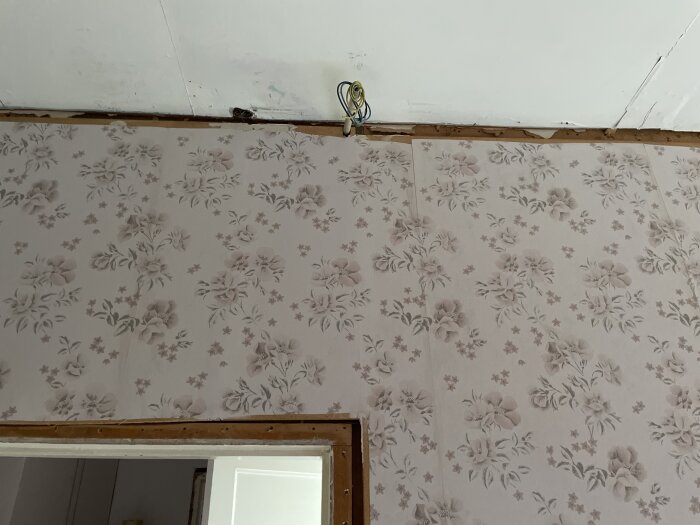 Vägg med blommig tapet och ett VP-rör med kablar synligt under ett vitt innertak innan regelarbetet i sovrum.