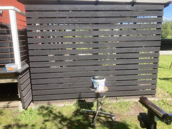 Svart trävägg byggd som vindskydd med burk målarfärg på en pall framför.