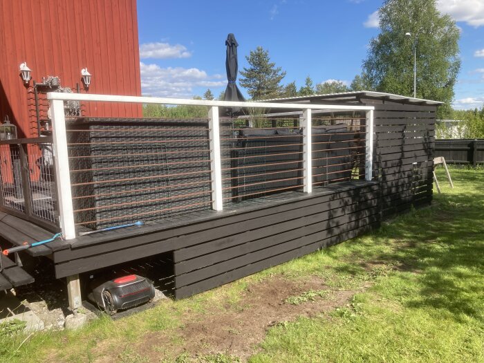 Svartmålat trädäck mot en röd vägg med svart skärmtak. En robotgräsklippare syns under trädäcket.