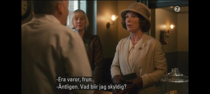 Kvinnor i gammaldags kläder står i en butik och pratar med en man. Texter på skärmen säger "Era varor, frun" och "Äntligen. Vad blir jag skyldig?".