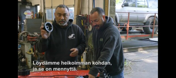 Två personer i verkstad, en visar upp en del av en maskin. En SUV syns i bakgrunden, och finsk text visas på skärmen.
