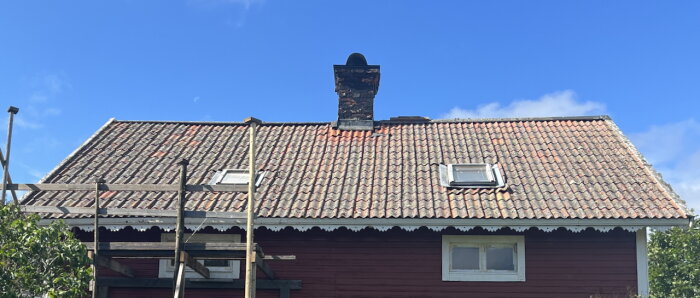 Rött sommarhus från 1800-talet med tegeltak och skorsten, två takfönster och byggställning på ena sidan.