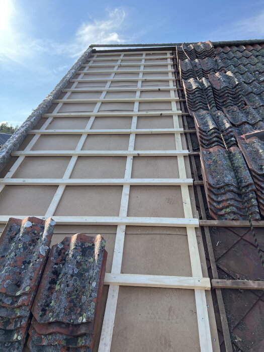 Takrenovering på ett sommarhus med tegelpannor delvis borttagna, visar träkonstruktion och masonit-diamanter under klart blå himmel.