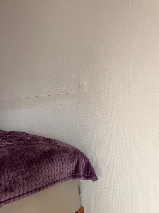 Vägg med diagonalt mönstrad tapet, delvis överlappande, med en säng täckt av lila filt i nedre delen av bilden.