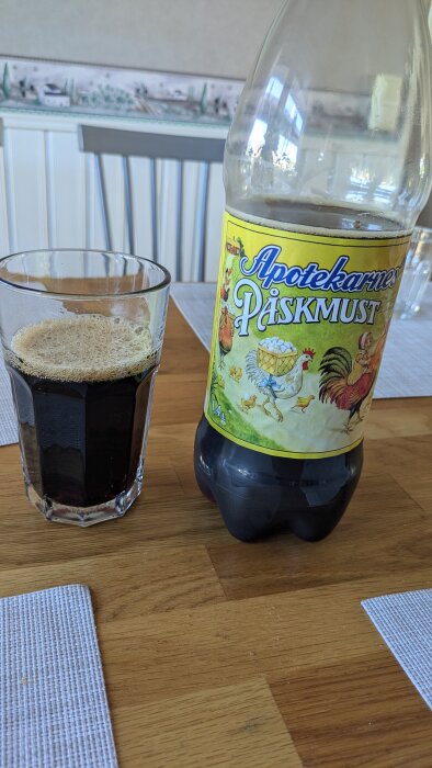 En flaska Apotekarnes Påskmust och ett glas av drycken på ett träbord.