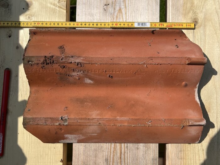 Tegelpanna från Heby tegelverk visas på en träyta, märkt med måttband och penna.