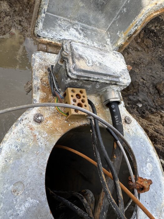 Öppen borrbrunn med metalllock, elkablar, och kondens på ytan. Flera kablar syns inuti brunnen.
