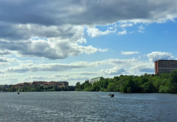 Sjölandskap med motorbåt på vattnet, omgiven av träd med byggnader i bakgrunden och molnig himmel.