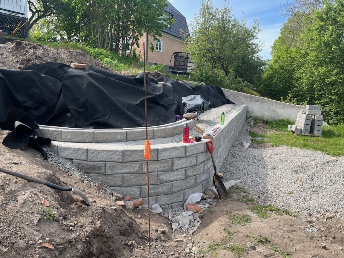 Mur av Benders sten byggs i trädgård, delvis täckt med svart duk. Spade och byggmaterial syns. Fråga om billigare toppstenar nämnd.